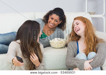 Cheerful young female friends with remote control and popcorn bowl on sofa at home
