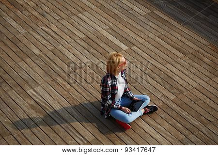 Young tourist sitting on a wooden floor with a tablet in hand enjoying rest and looks aside