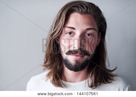 Young caucasian man with long brwon hair hanging loose with a beard, wearing a whire t-shirt standing in front of a white wall