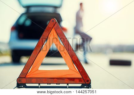 A young man with a silver car that broke down on the road.He has set up a warning triangle.He is waiting for the technician to arrive.