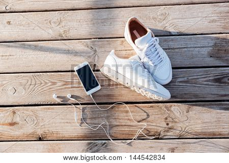 Sport concept - pair of white sneakers, smartphone and earphones over wooden background