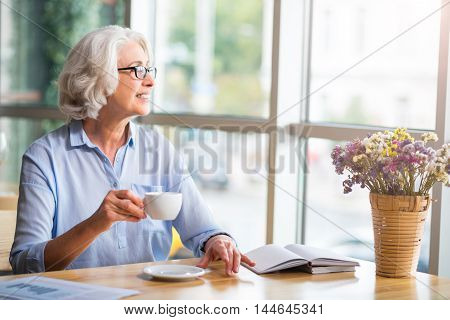 Enjoy every day. Cheerful content smiling woman drinking coffee and looking in the window while resting at the table in the cafe