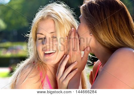 Two beautiful blond and brunette women whispering to each other outside in sunny weather