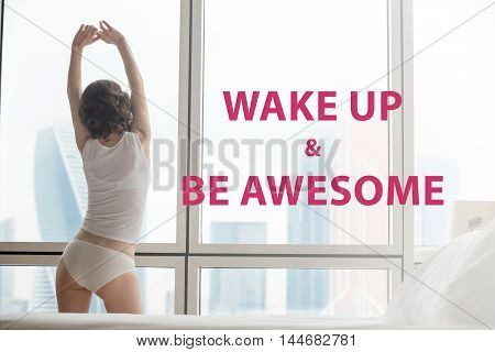 Back view of young woman stretching after waking up doing morning exercises at home. Female model relaxing and looking at misty city scenery in the window. Motivational phrase 