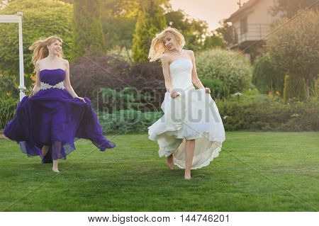 Bride and bridesmaid outside. Two beautiful girls running on lown. Bride in wedding dress. Bridesmaid in a purple evening gown. Hair fluttering in the wind
