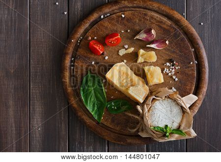 Cheese delikatessen closeup on rustic wood. Wooden desk with parmesan, camembert and brie cuts decorated with garlic, tomato and basil, top view image with copy space