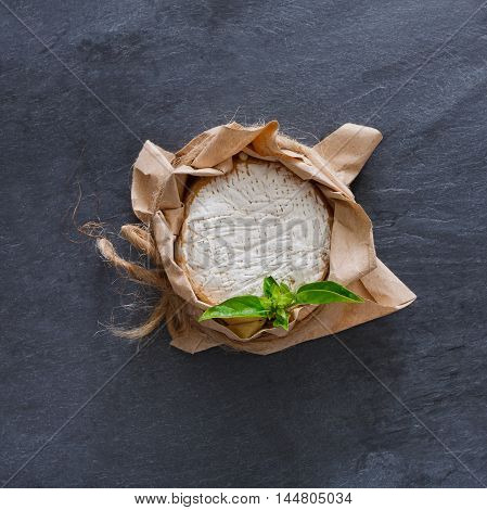 Cheese delikatessen closeup on black stone surface. Camembert or brie circle in brown kraft paper decorated with basil, top view image