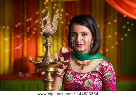 Indian young and beautiful girl in traditional wear lighting oil lamp or samai with diya and celebrating ganesh festival or Diwali or deepavali. Indian girl hands holding oil lamp indoors.