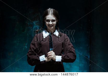 Portrait of a young girl with knife in school uniform as killer woman against school board . The image in the style of Halloween and Addams family