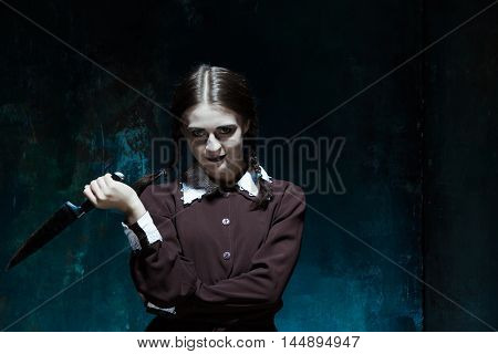 Portrait of a young girl with knife in school uniform as killer woman against school board . The image in the style of Halloween and Addams family