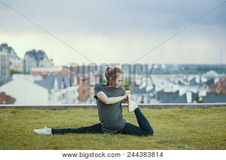 Young Female Acrobat On The Grass Performs Twine, Cityscape On Background, Horizontal