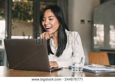 Photo of cheerful asian woman 20s wearing white shirt laughing and pointing finger at screen of laptop while speaking or chatting on video call in office