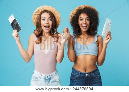 Photo of caucasian and african american women 20s wearing straw hats smiling at camera and holding passports with travel tickets isolated over blue background