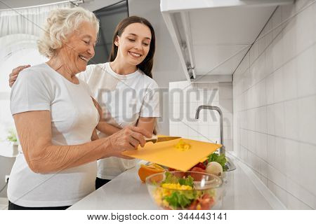 Side View Of Young Brunette Girl Hugging Grandmother In White Kitchen. Smiling Senior Woman Throwing