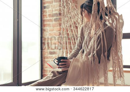 Beautiful young woman, which is wearing cashmere nightwear clothes, drinking tea or coffee and reading book while relaxing on hammock in scandinavian cabin near big window