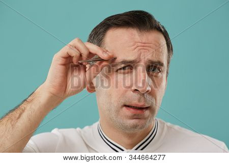 Head And Shoulders Portrait Of Wincing Middle Aged Man Plucking Eyebrows Standing Against Blue Backg