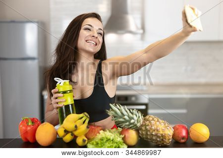 Fit Young Woman Wearing Sexy Black Sports Top Shooting Selfie Standing In The Kitchen Full Of Fruits