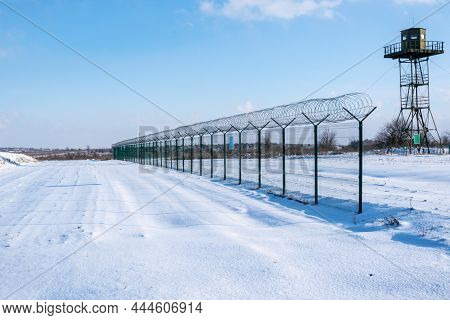 Border Fence With Barbed Wire In Front Of The Surveillance Tower In A Snow-covered Field Against A B