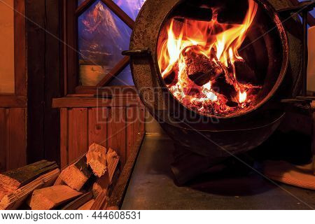 A Fire Is Burning In The Firebox Of A Round Cast-iron Stove With An Open Door. Firewood Is Lying Nea
