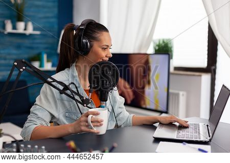 Woman Vlogger Holding Cup And Streaming Podcast Using Laptop. Influencer With Headphones In Home Off