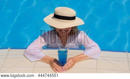 Happy Senior Woman Relaxing Near Blue Outdoor Swimming Pool With Blue Cocktail Wearing Straw Hat. Pe