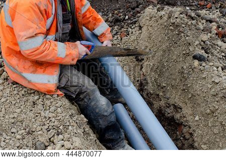 Groundworker In Hi-viz Coat Cutting Grey Plastic Telecom Duct With Hand Saw While Sitting At The Edg