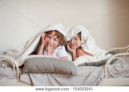 Two children brother and sister squirmy on the bed in the bedroom. They look out from under a blanket and laugh.