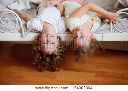 The children boy and girl naughty on the bed in the bedroom. They lie side by side their heads hanging off the bed. Children is very amused.