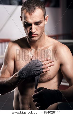 In stepwise fashion. Handsome muscular man standing in the gym and carefully wrapping his hand with grey boxing tape.