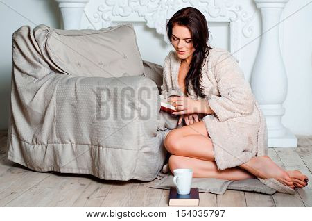 attractive young brunette woman at home beige bathrobe, sitting on the wooden floor Aper on an easy chair and reading a book at home.