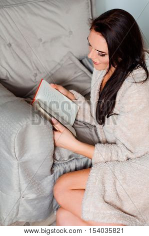 attractive young brunette woman at home beige bathrobe, sitting on the wooden floor Aper on an easy chair and reading a book at home.