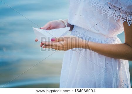 Young girl in a white dress holding a white paper boat on a background of blue sea. Summer by the sea