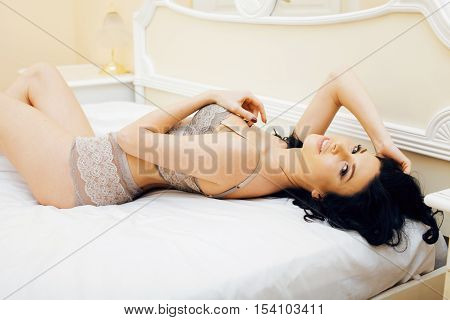 Sexy beautiful brunette woman lying in bed sensual gray lingerie, looking at camera. Seduction concept in luxury room interior close up