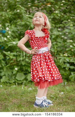Little girl in red polka-dotted dress stands with  her hands akimbo and looking upward at grassy lawn.