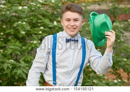Smiling teenage boy with green hat in hand against bushes on summer day.
