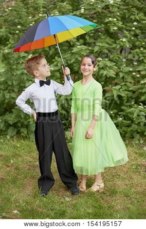 Little boy and girl dressed in dance suits stand outdoor under rainbow coloured umbrella.