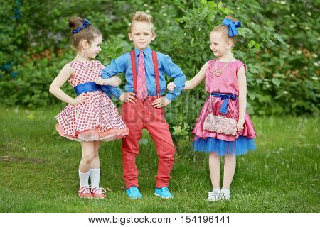 Boy in red trousers and blue shirt stands with two girls at grassy lawn.