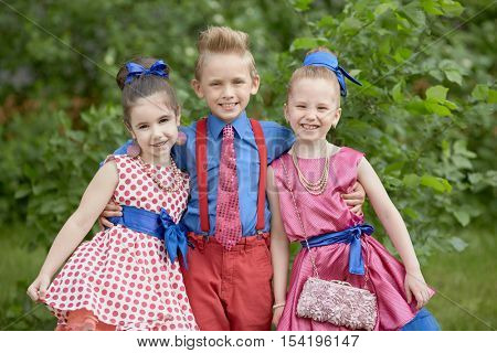 Boy in red trousers and blue shirt stands embracing two girls at grassy lawn.