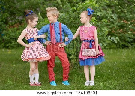 Boy in red trousers and blue shirt stands with two girls at grassy lawn.