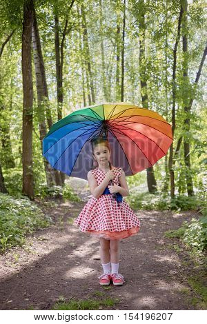 Little girl in polka-dotted dress stands with rainbow coloured umbrella in summer park.