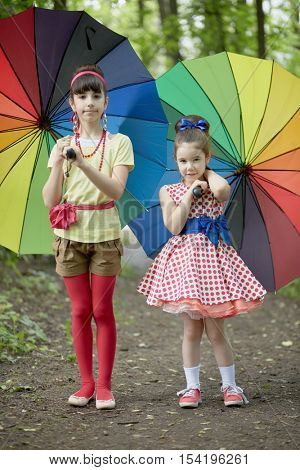 Two girls stands with rainbow coloured umbrellas in summer park.