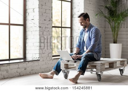 Online interaction. Happy pleasant bearded man typing a message and looking at the screen of his laptop while communicating via the Internet