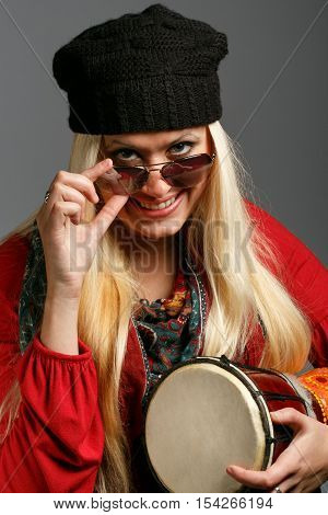 Young beautiful smiling blonde woman in colorful clothes black hat and sunglasses with a cheerful look holding small ethnic djembe drum at grey background