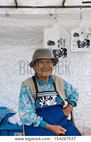 Takayama Japan - September 24 2016: Closeup of old smiling lady who is a vendor on the public morning market in front of the Jinya Hall. Blue clothes white tarp background.