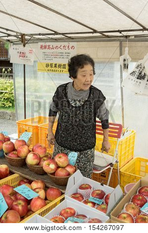 Takayama Japan - September 24 2016: Older active lady who is a vendor on the public morning market in front of the Jinya Hall promotes her red apples. Black clothes white tarp background.