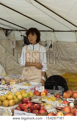 Takayama Japan - September 24 2016: Middle aged lady who is a vendor on the public morning market in front of the Jinya Hall promotes her red and yellow apples. Beige clothes white tarp background.