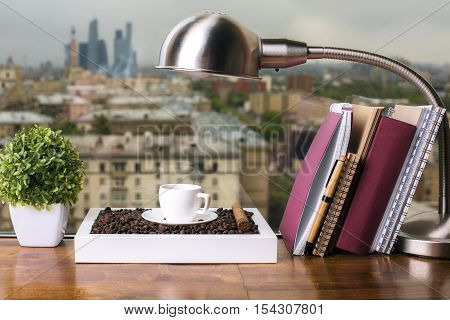 Closeup of wooden windowsill with coffee cup and beans table lamp decorative plant and book with notepads on city background