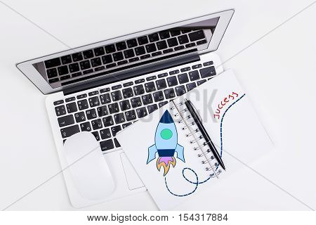 Top view of white desktop with laptop computer mouse and spiral notepad with creative rocket drawing. Business start up concept. Mock up