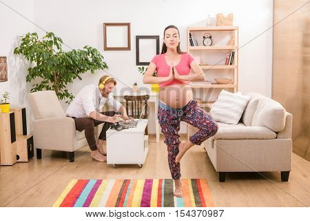 Health and pregnancy concept. Young pregnant woman training yoga at home. Brunette lady clasped her hands and concentrating on baby. Her husband using laptop computer for playing compuer games.