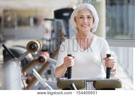 Cheerful workout. Attractive senior joyful woman smiling and staying fit by using equipment in a gym.